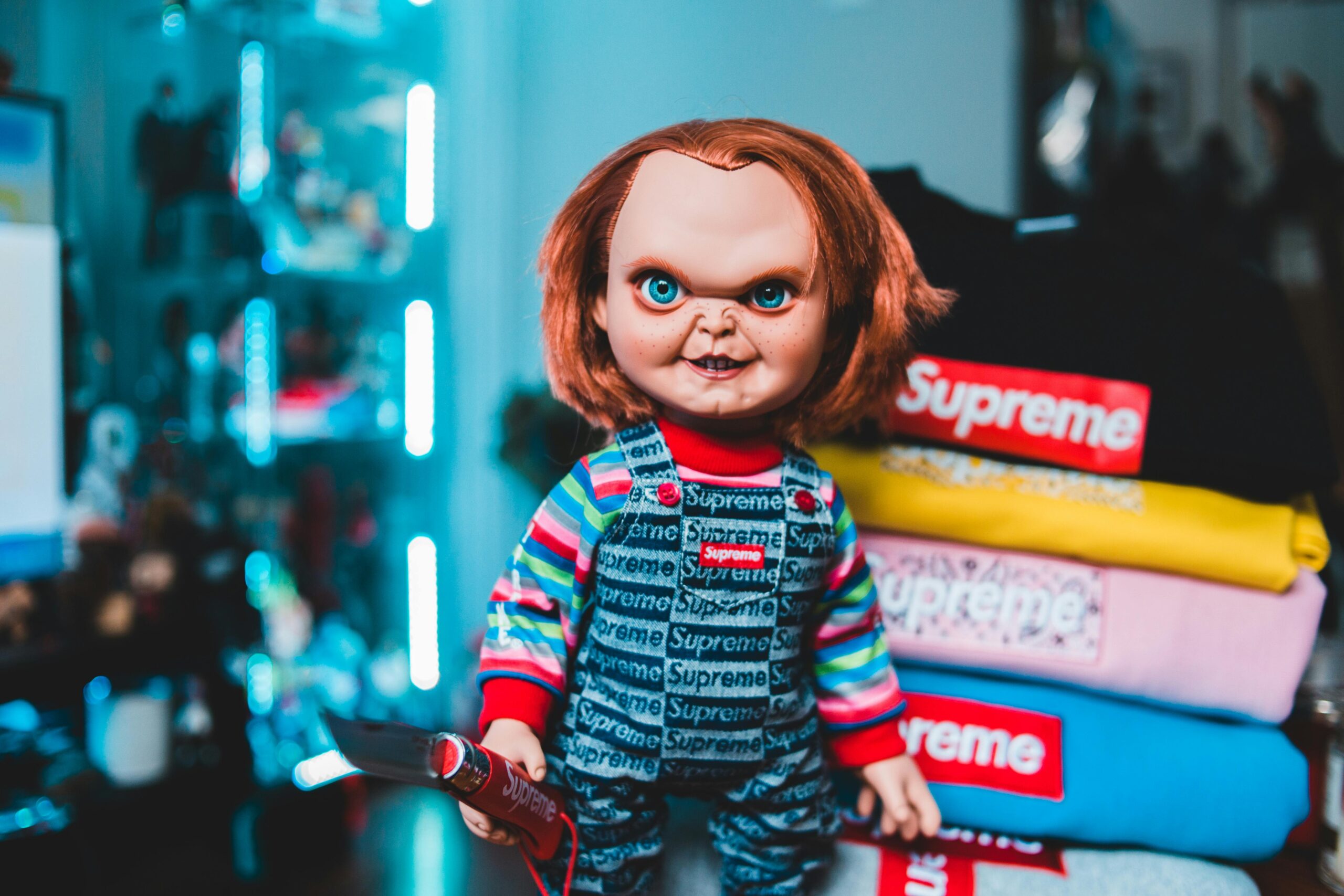 Scary Chucky doll wearing a Supreme outfit stands next to colorful Supreme clothing indoors.